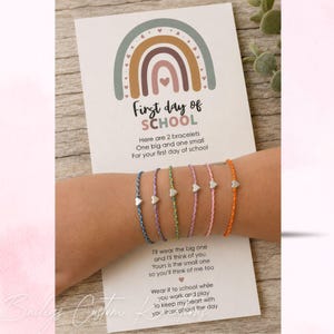 Peut inclure: Un ensemble de cinq bracelets en cordon fin bleu, violet, vert, rose et orange, chacun orné d'un petit cœur en argent. Les bracelets sont présentés sur une carte blanche avec un motif arc-en-ciel et le texte First day of SCHOOL.