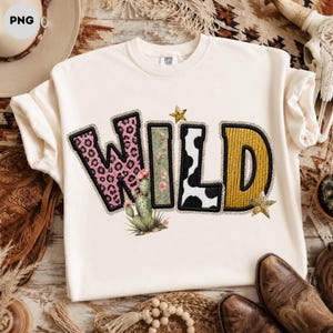 May include: A cream-colored crewneck sweatshirt featuring the word WILD in large, patterned letters. The W has a pink leopard print, the I is green with floral details, the L is black and white cow print, and the D is textured mustard yellow.