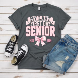 May include: A gray t-shirt with the text "MY LAST FIRST DAY SENIOR 2026" in pink. The shirt is paired with denim shorts and black canvas sneakers with white soles and laces. The items are arranged on a light wood-grain surface.
