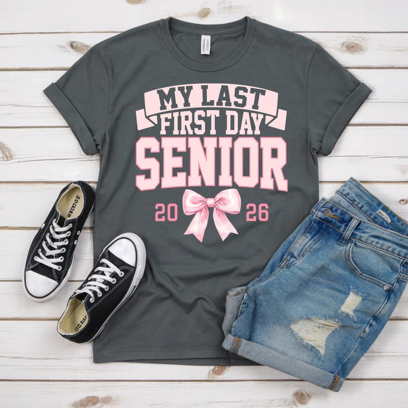 May include: A gray t-shirt with the text "MY LAST FIRST DAY SENIOR 2026" in pink. The shirt is paired with denim shorts and black canvas sneakers with white soles and laces. The items are arranged on a light wood-grain surface.