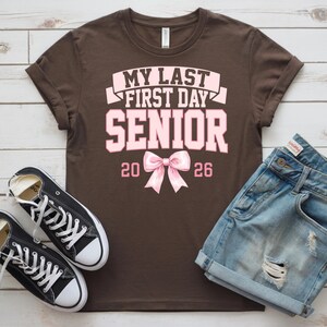 May include: A brown t-shirt with the text "MY LAST FIRST DAY SENIOR 2026" in pink. The shirt is paired with denim shorts and black canvas sneakers with white laces.