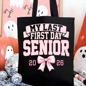 May include: Black tote bag with pink text that reads "MY LAST FIRST DAY SENIOR 2026". The bag has two black handles and a pink bow graphic. The background includes Halloween decorations like pumpkins and disco balls.