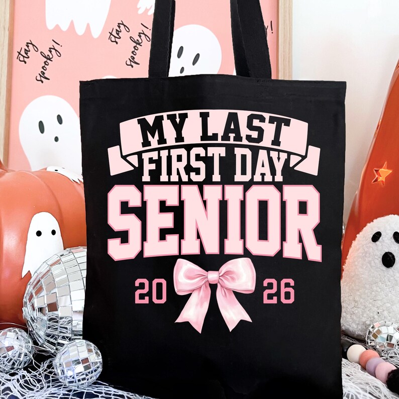 May include: Black tote bag with pink text that reads "MY LAST FIRST DAY SENIOR 2026". The bag has two black handles and a pink bow graphic. The background includes Halloween decorations like pumpkins and disco balls.