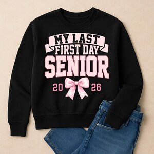 May include: Black sweatshirt with the text "MY LAST FIRST DAY SENIOR 2026" in pink. A pink bow graphic is centered below the text. Blue jeans are partially visible in the image.