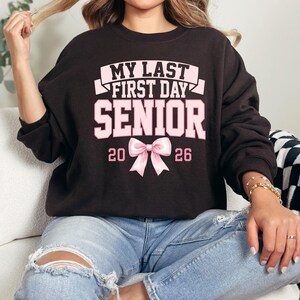 May include: A dark brown sweatshirt with the text "MY LAST FIRST DAY SENIOR 2026" in white and pink. A pink bow graphic is centered below the word "SENIOR". The sweatshirt is worn with blue jeans.