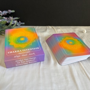 May include: A deck of Chakra Intention oracle cards in a rectangular box and a matching stack of cards. The box and cards feature a colorful gradient background with a central mandala design and the text: Chakra Intention, Align Heal Awake.