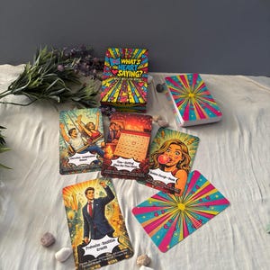 May include: A deck of What's The Heart Saying? oracle cards featuring vibrant pop art illustrations. The set includes a box, a stack of cards, and several individual cards displaying scenes like a person in a suit, a person blowing a bubble, and a couple on a swing, all with colorful sunburst patterns.