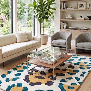 May include: A modern living room featuring a cream-colored sofa, two gray armchairs, and a rectangular coffee table with a glass top. A patterned rug with blue, gold, and black shapes sits on the wooden floor. A large potted plant and a bookshelf complete the scene.