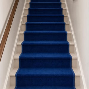 Op de afbeelding: Een trap met witte treden en een diepblauwe tapijtloper. De loper bedekt het midden van de treden, met een bijpassende blauwe rand. Een houten leuning loopt langs de linkerkant. Een ingelijste foto hangt aan de muur.