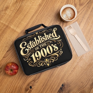 Może przedstawiać: Czarna torba na lunch ze złotym napisem "Established in the 1900's". Torba ma czarny uchwyt i zamek błyskawiczny. Jabłko, filiżanka kawy i drewniany widelec leżą na drewnianej powierzchni.
