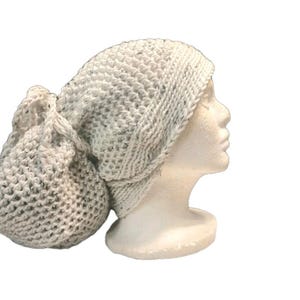 Könnte beinhalten: Eine hellgraue Strickmütze mit passendem Beutel. Die Mütze ist aus einem grob gestrickten Material gefertigt und wird auf einem weißen Schaufensterkopf präsentiert. Der Beutel ist rund und hat einen Kordelzug.