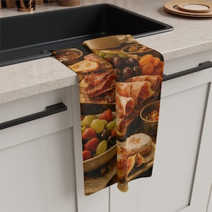 May include: A kitchen towel featuring a printed image of a charcuterie board with various food items. The towel displays a bowl of fruit, cured meats, cheese, and other delicacies. The color palette includes browns, reds, and greens.