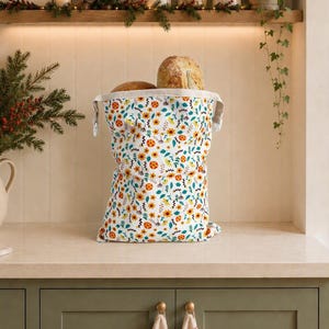 May include: A fabric bread bag featuring a repeating pattern of orange flowers, green leaves, and small dark blue geometric shapes, containing two round loaves of bread, positioned on a countertop.
