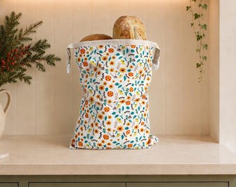 Könnte beinhalten: Ein Stoffbeutel für Brot mit einem wiederkehrenden Muster aus orangefarbenen Blumen, grünen Blättern und kleinen dunkelblauen geometrischen Formen, der zwei runde Brotlaibe enthält und auf einer Arbeitsplatte steht.