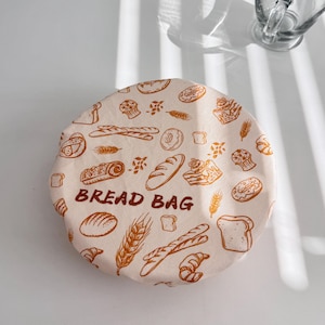 Op de afbeelding: Een beige broodzak met een bruin broodthema-patroon en de woorden "BREAD BAG" in bruin gedrukt. De ronde zak ligt op een wit oppervlak, met een doorzichtige glazen mok op de achtergrond.