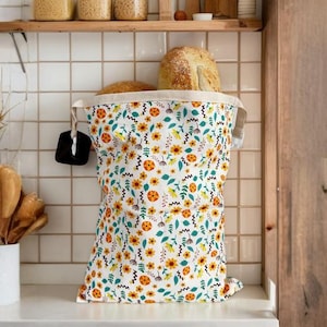May include: A white bread bag with a floral pattern of orange, yellow, and blue flowers. The bag is open and filled with bread. The bag is on a white countertop in a kitchen setting.