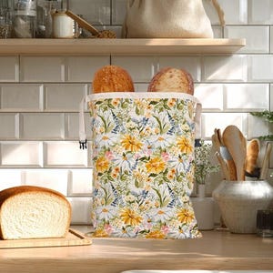 May include: A loaf of sliced bread on a wooden board beside a floral bread bag filled with bread. The bag has a white background with yellow and white flowers and green leaves. A beige bag sits on a shelf.
