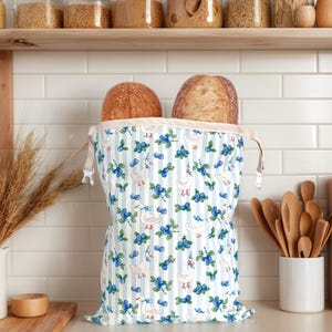 May include: A bread bag with a blue and white striped pattern, featuring illustrations of geese and blueberries. Two loaves of bread are visible inside the bag. The bag is on a kitchen counter with wooden utensils and jars in the background.