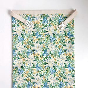 Op de afbeelding: Een rechthoekig stoffen item met een bloemenpatroon in groen, blauw en geel, met de tekst "Green Flowers" in blauw bovenaan. Het heeft een beige band met een witte plastic gesp.