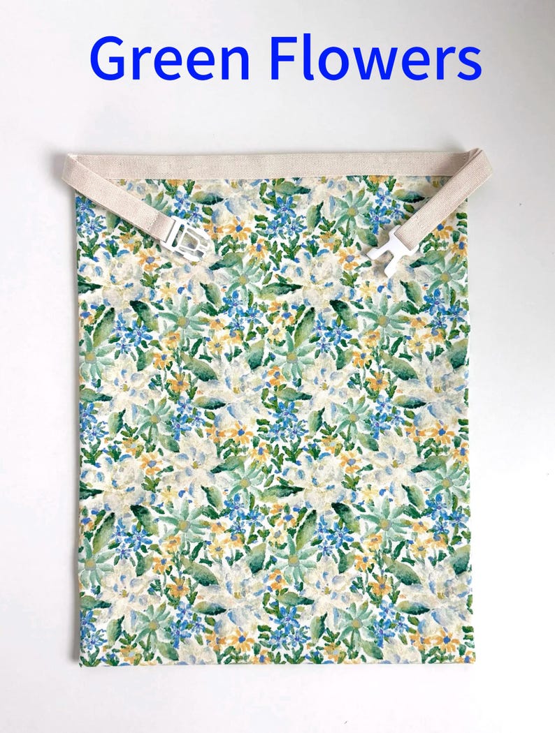 Op de afbeelding: Een rechthoekig stoffen item met een bloemenpatroon in groen, blauw en geel, met de tekst "Green Flowers" in blauw bovenaan. Het heeft een beige band met een witte plastic gesp.