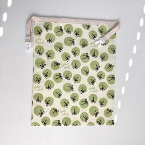 Op de afbeelding: Een rechthoekige trekkoordtas met een herhalend patroon van groene appelillustraties op een crèmekleurige achtergrond. De tas heeft een beige rand en een beige trekkoord met een witte plastic sluiting. De woorden "Green Apple" staan op de tas.