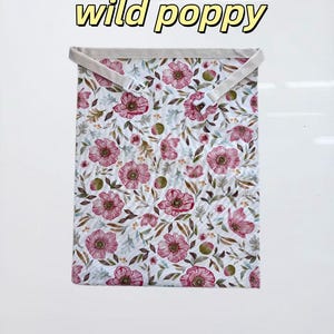 Peut inclure: Un sac en tissu rectangulaire avec un motif floral de coquelicots roses et de feuilles vertes sur fond blanc. Le texte "wild poppy" est imprim&eacute; au-dessus du sac en lettres jaunes avec un contour noir. Il comprend une sangle beige avec une boucle en plastique blanc.