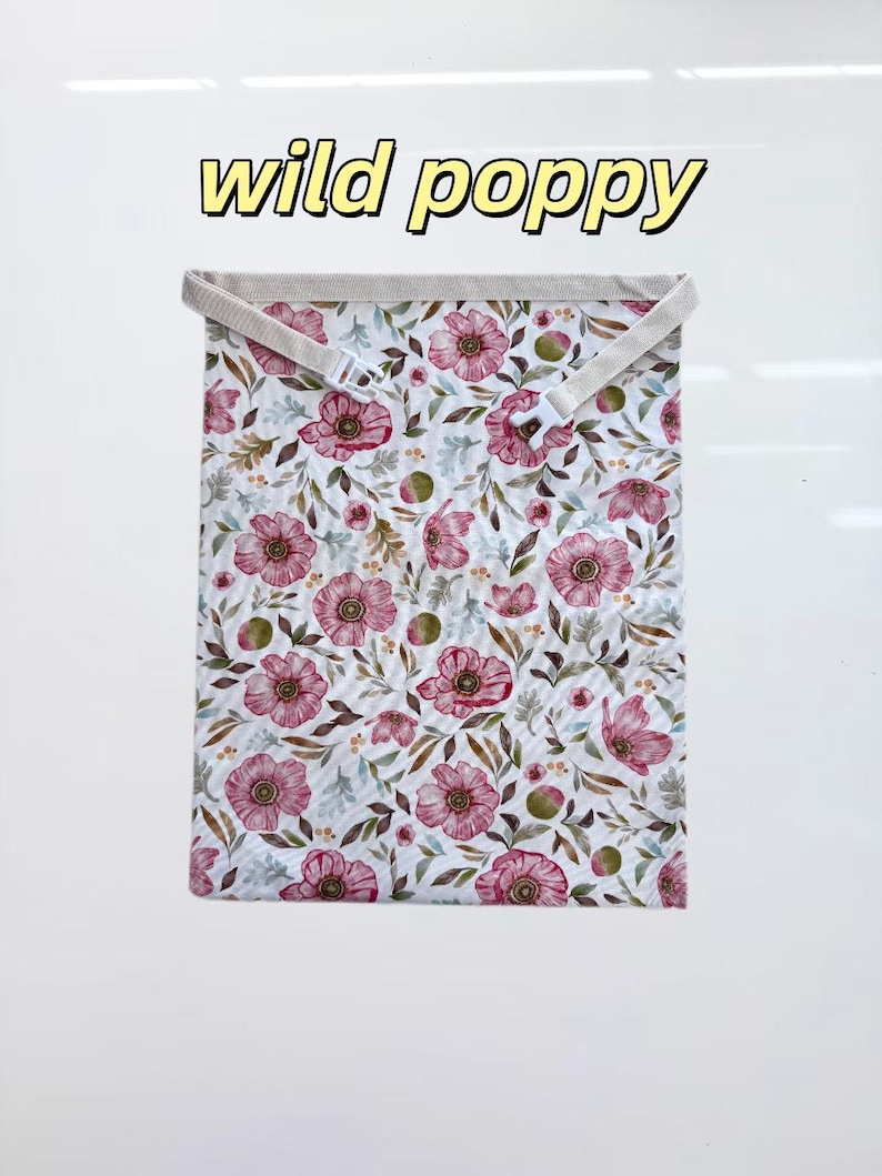 Peut inclure: Un sac en tissu rectangulaire avec un motif floral de coquelicots roses et de feuilles vertes sur fond blanc. Le texte "wild poppy" est imprim&eacute; au-dessus du sac en lettres jaunes avec un contour noir. Il comprend une sangle beige avec une boucle en plastique blanc.