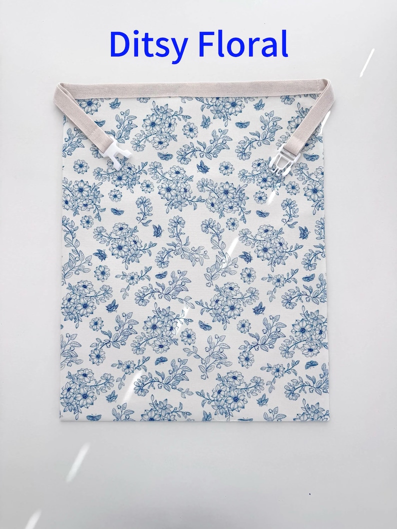 Peut inclure: Un sac en tissu rectangulaire avec un motif floral bleu sur fond blanc. Le bord sup&eacute;rieur comporte une sangle beige avec deux boucles en plastique blanc. Le texte Ditsy Floral est imprim&eacute; en bleu au-dessus du sac.