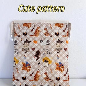 Op de afbeelding: Een beige trekkoordtas met een herhalend patroon van cartoonkatten met heksenhoeden, ketels en pompoenen. De tekst "Cute pattern" wordt in het geel weergegeven. De woorden "Magic Cat" zijn ook zichtbaar.