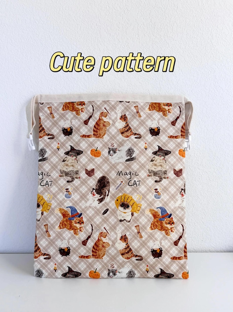 Op de afbeelding: Een beige trekkoordtas met een herhalend patroon van cartoonkatten met heksenhoeden, ketels en pompoenen. De tekst "Cute pattern" wordt in het geel weergegeven. De woorden "Magic Cat" zijn ook zichtbaar.