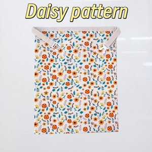 Op de afbeelding: Een rechthoekig stoffen item met een madeliefjespatroon, met gele, oranje en blauwgroene bloemen op een witte achtergrond. De tekst "Daisy pattern" wordt in een gele lettertype weergegeven. Het item heeft twee beige bandjes met zilveren gespen.