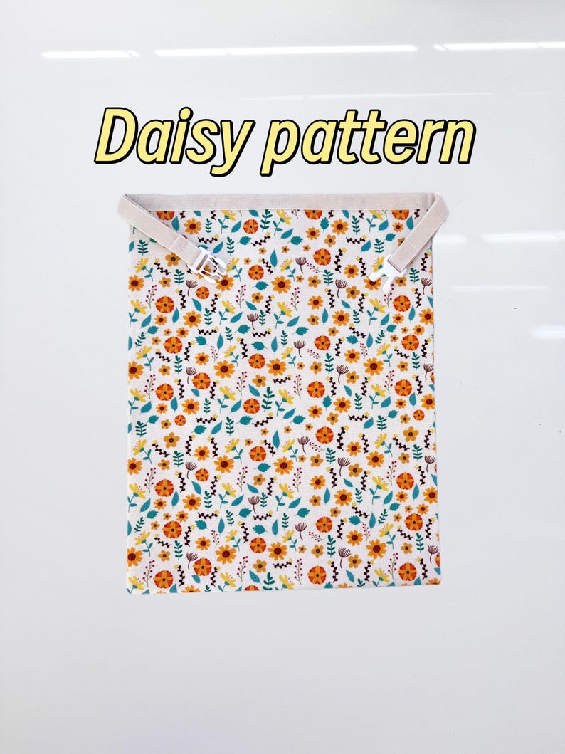 Op de afbeelding: Een rechthoekig stoffen item met een madeliefjespatroon, met gele, oranje en blauwgroene bloemen op een witte achtergrond. De tekst "Daisy pattern" wordt in een gele lettertype weergegeven. Het item heeft twee beige bandjes met zilveren gespen.