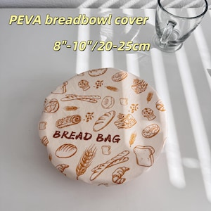 Op de afbeelding: Een beige PEVA broodkomhoes met een broodthema, inclusief afbeeldingen van brood, gebak en tarwe. De hoes is voorzien van het opschrift "BREAD BAG" en is geschikt voor kommen met een diameter van 20-25cm.