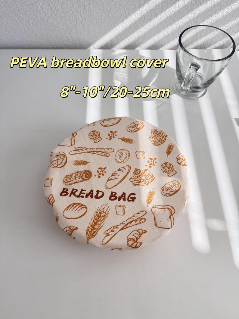 Op de afbeelding: Een beige PEVA broodkomhoes met een broodthema, inclusief afbeeldingen van brood, gebak en tarwe. De hoes is voorzien van het opschrift "BREAD BAG" en is geschikt voor kommen met een diameter van 20-25cm.