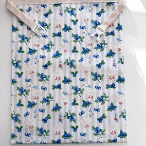 Op de afbeelding: Een stoffen tas met een herhalend patroon van witte eenden met feesthoeden en takjes bosbessen op een lichtblauwe en wit gestreepte achtergrond. De tas heeft een beige band met een witte plastic gesp. De tekst "Blueberry Duck" staat bovenaan.