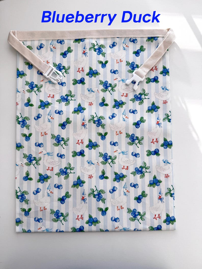 Op de afbeelding: Een stoffen tas met een herhalend patroon van witte eenden met feesthoeden en takjes bosbessen op een lichtblauwe en wit gestreepte achtergrond. De tas heeft een beige band met een witte plastic gesp. De tekst "Blueberry Duck" staat bovenaan.
