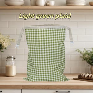 Peut inclure: Un sac en tissu rectangulaire avec un motif &agrave; carreaux vert clair et blanc. Le sac pr&eacute;sente une large ouverture sup&eacute;rieure de couleur claire avec deux sangles lat&eacute;rales et des clips en plastique blanc. Le texte "Light green plaid" est inscrit au-dessus.