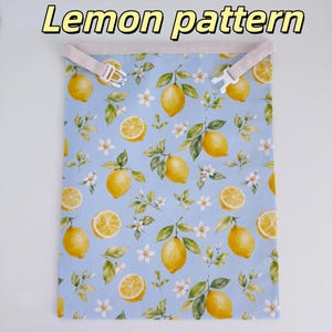 Peut inclure: Un sac en tissu rectangulaire avec un fond bleu clair orn&eacute; d'un motif r&eacute;p&eacute;titif de citrons jaunes, de feuilles vertes et de fleurs blanches. Le bord sup&eacute;rieur comporte une sangle beige avec une boucle en plastique blanc. Texte Lemon pattern.