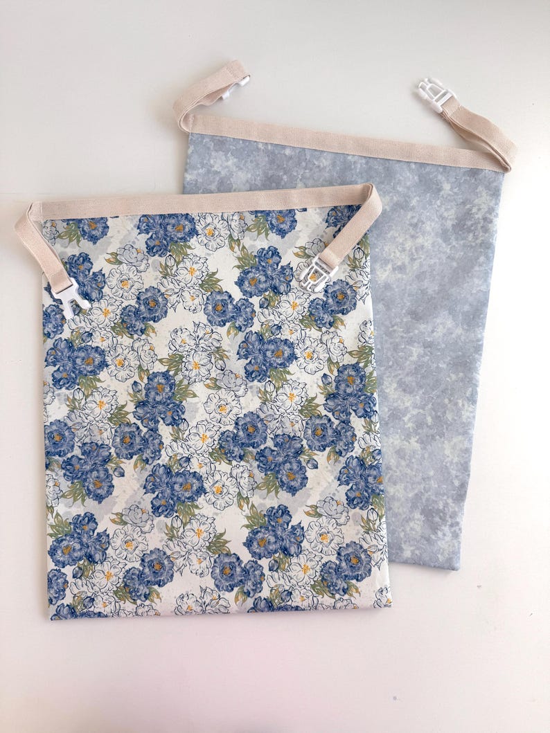 Op de afbeelding: Twee stoffen tassen met beige bandjes en plastic gespen. E&eacute;n tas heeft een bloemenpatroon met blauwe en witte bloemen op een cr&egrave;mekleurige achtergrond. De andere tas heeft een lichtblauw, gemarmerd patroon.
