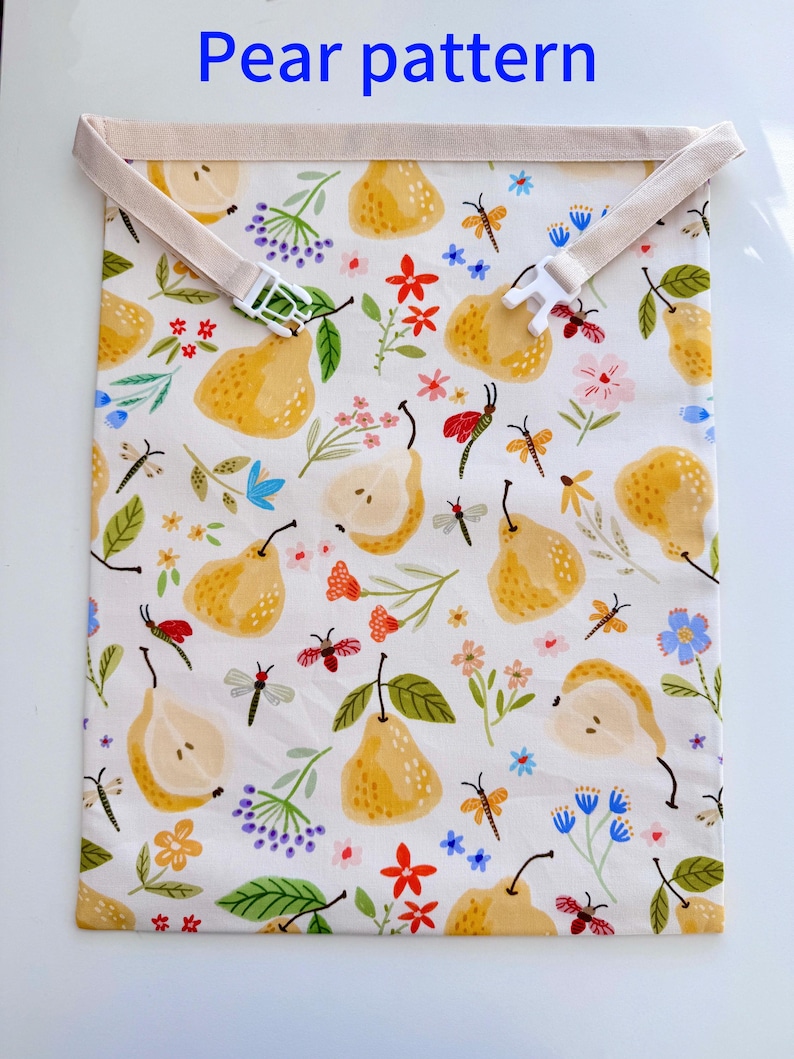 Op de afbeelding: Witte stoffen tas met een perenpatroon, met gele peren, kleurrijke bloemen en insecten. De tas heeft een beige band met een witte plastic gesp. De tekst "Pear pattern" staat bovenaan.