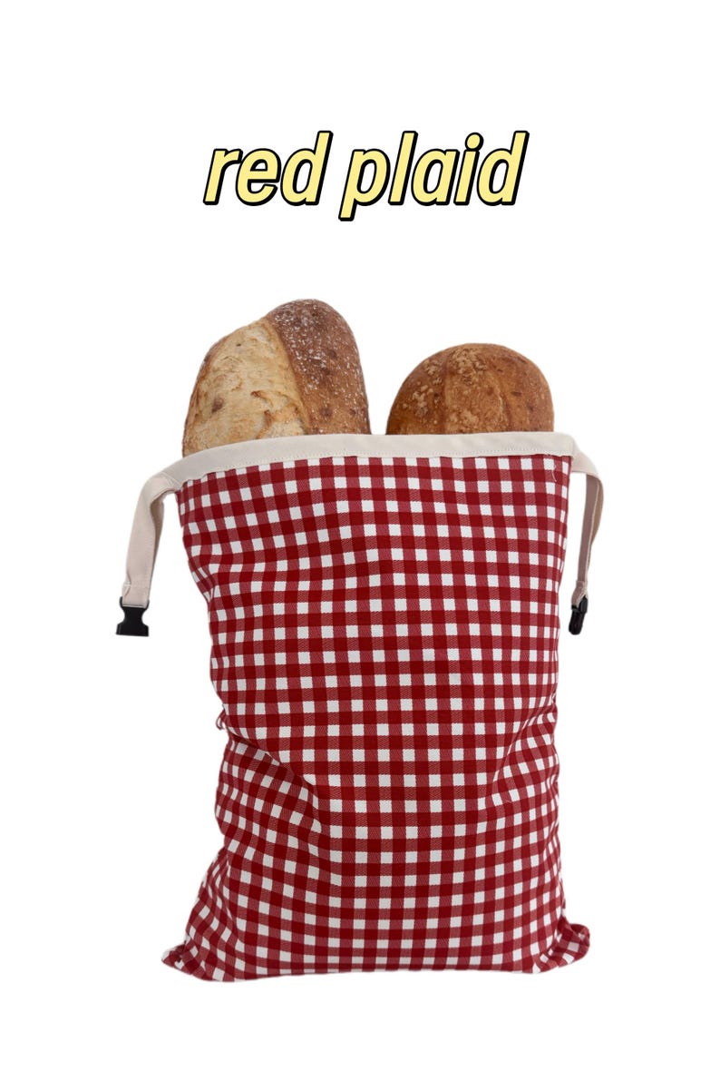 Peut inclure: Un sac &agrave; pain &agrave; motif vichy rouge et blanc avec une bordure sup&eacute;rieure cr&egrave;me et des boucles lat&eacute;rales en plastique noir. Deux miches de pain croustillantes sont visibles &agrave; l'int&eacute;rieur. Le texte "red plaid" est &eacute;crit en jaune au-dessus.