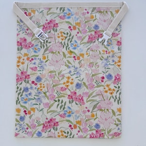 Op de afbeelding: Een bloemenpatroon tas met de tekst "Pink Flowers" bovenaan. De tas heeft een cr&egrave;mekleurige achtergrond met roze, blauwe en gele bloemen. Het heeft een cr&egrave;mekleurige band met een witte plastic gesp.