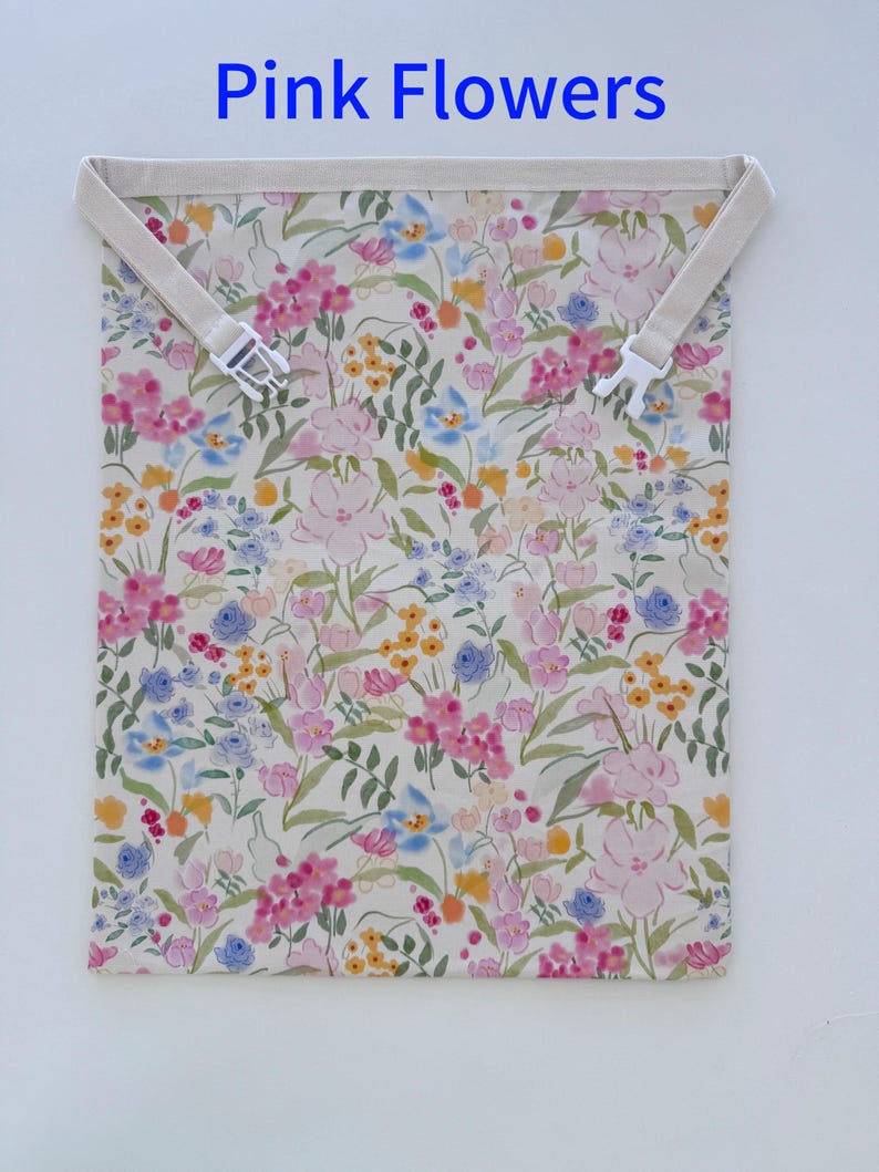 Op de afbeelding: Een bloemenpatroon tas met de tekst "Pink Flowers" bovenaan. De tas heeft een cr&egrave;mekleurige achtergrond met roze, blauwe en gele bloemen. Het heeft een cr&egrave;mekleurige band met een witte plastic gesp.