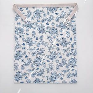 Op de afbeelding: Een wit stoffen item met een blauw bloemenpatroon en de tekst "Ditsy Floral" in blauw. Het heeft een beige band met een witte plastic clip. Het bloemendesign omvat bloemen en vlinders.