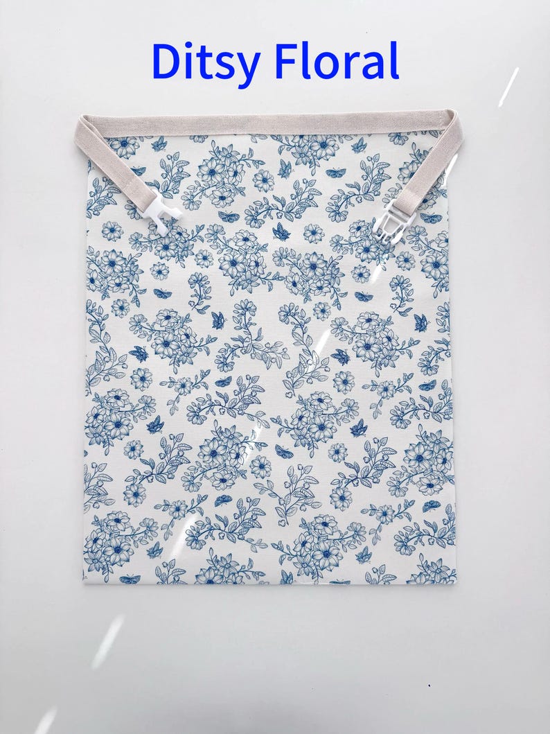 Op de afbeelding: Een wit stoffen item met een blauw bloemenpatroon en de tekst "Ditsy Floral" in blauw. Het heeft een beige band met een witte plastic clip. Het bloemendesign omvat bloemen en vlinders.