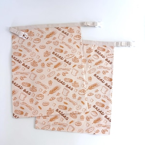 Puede incluir: Dos bolsas de pan beige con un patrón repetido de ilustraciones de pan marrón. Las bolsas tienen un cierre de cordón y las palabras "BREAD BAG" están impresas en ellas. Las bolsas están hechas de una tela suave y duradera.