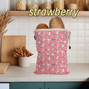 Op de afbeelding: Een roze broodzak met aardbeienpatroon, gevuld met twee broden. De tas heeft een trekkoordsluiting en een witte binnenkant. Het woord "strawberry" staat in het geel boven de tas.