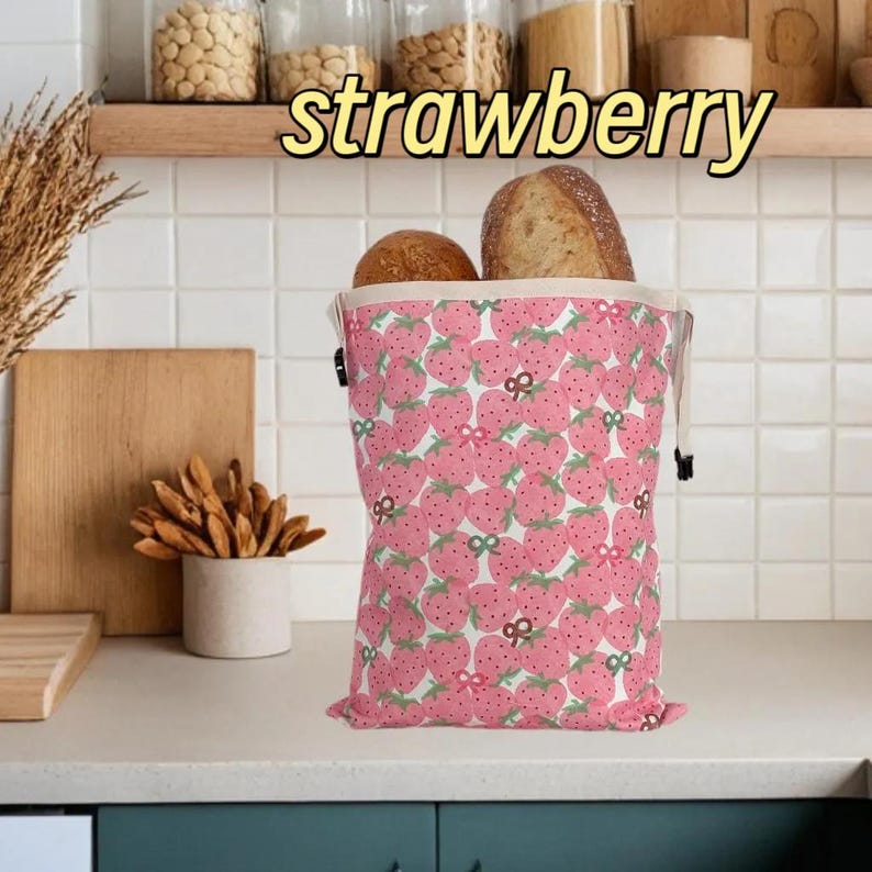 Op de afbeelding: Een roze broodzak met aardbeienpatroon, gevuld met twee broden. De tas heeft een trekkoordsluiting en een witte binnenkant. Het woord "strawberry" staat in het geel boven de tas.