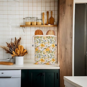 Op de afbeelding: Een bloemenbroodzak, gevuld met twee broden, staat op een wit aanrecht. De zak heeft een patroon van gele madeliefjes, blauwe bloemen en groene bladeren. De keukenomgeving omvat een houten plank met potten en keukengerei.