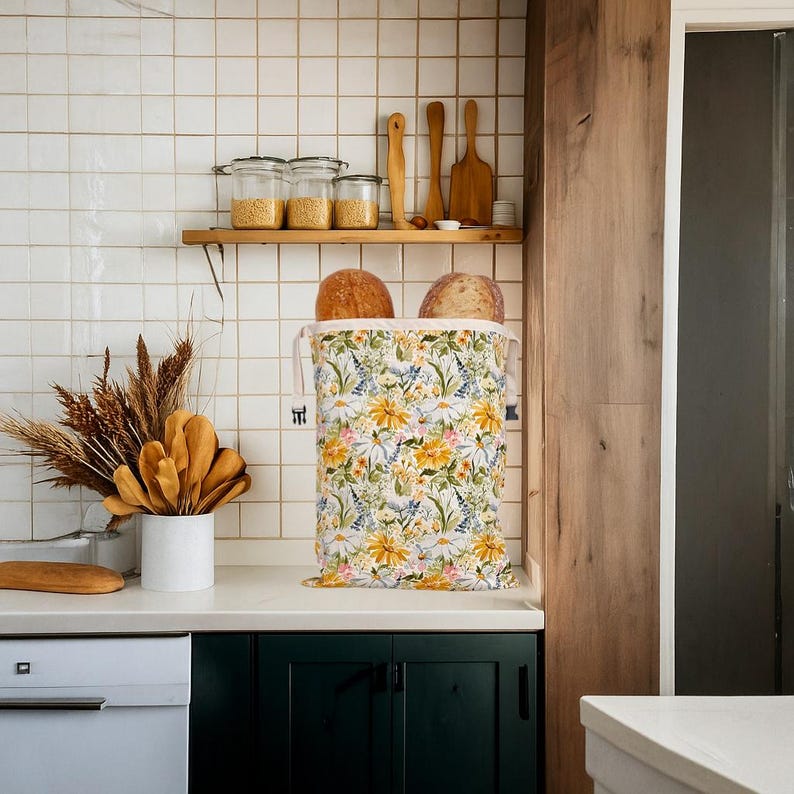 Op de afbeelding: Een bloemenbroodzak, gevuld met twee broden, staat op een wit aanrecht. De zak heeft een patroon van gele madeliefjes, blauwe bloemen en groene bladeren. De keukenomgeving omvat een houten plank met potten en keukengerei.